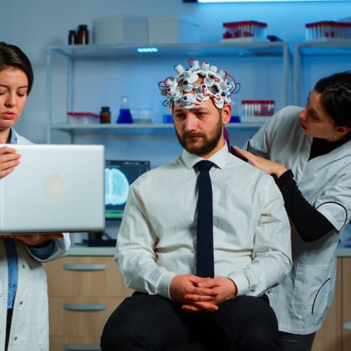 Medic in neuroscience working in neurological research laboratory developing brain experiment holding laptop explaning to man brainwave scanning headset side effects of nervous system treamtment.
